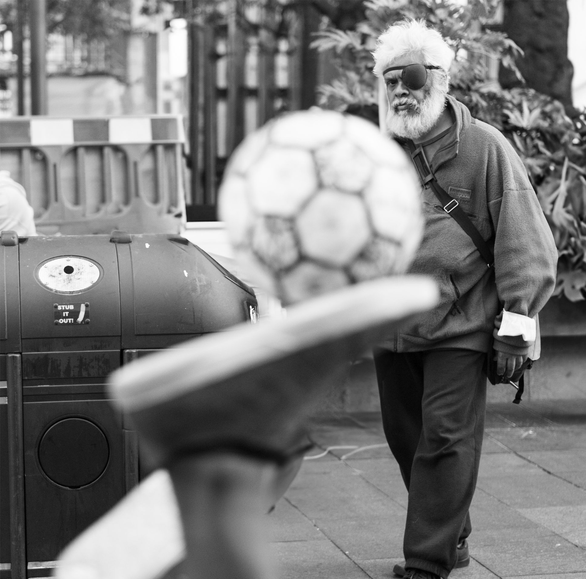 Man walking with a eyepatch on behind someone making football tricks in black and white